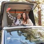 Mature multiracial friends taking a selfie on mini van camper roof top during road trip vacation