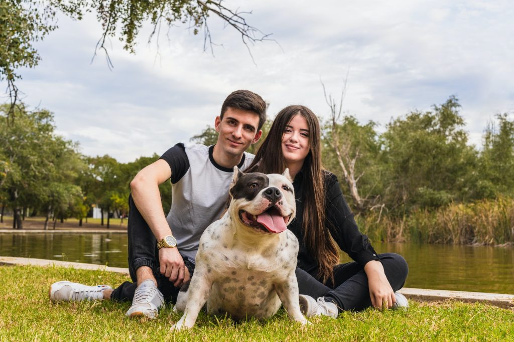 Two people sitting next to an american bully dog in a park next to a river