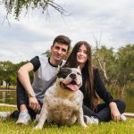 Two people sitting next to an american bully dog in a park next to a river
